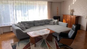 Ein Wohnzimmer mit grauem Ecksofa, einem kleinen Couchtisch, einem schwarzen Sessel, einer Holzkabine und einer Heizung.