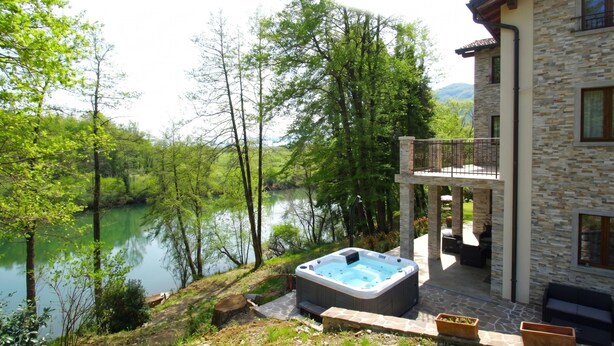 A stone building with a hot tub, outdoor seating, and a view of a lake surrounded by trees.