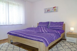 A bedroom with a wooden bed, purple bedding, a nightstand with a lamp, a white wall, and a window with sheer curtains.