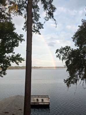 A dock with chairs and a table, surrounded by trees and a calm lake with a rainbow.