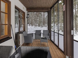 A covered outdoor seating area with wicker furniture, a glass table, and a view of a snowy forest.