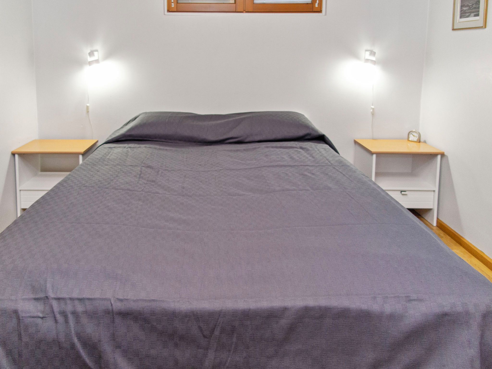 Ein Einzelbett mit grauer Bettdecke, zwei Nachttische mit Lampen und eine kleine Uhr auf einem der Tische.