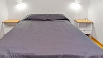 Ein Einzelbett mit grauer Bettdecke, zwei Nachttische mit Lampen und eine kleine Uhr auf einem der Tische.