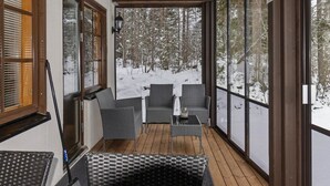 Een overdekt terras met rieten meubels, grote ramen en uitzicht op een besneeuwd bos.