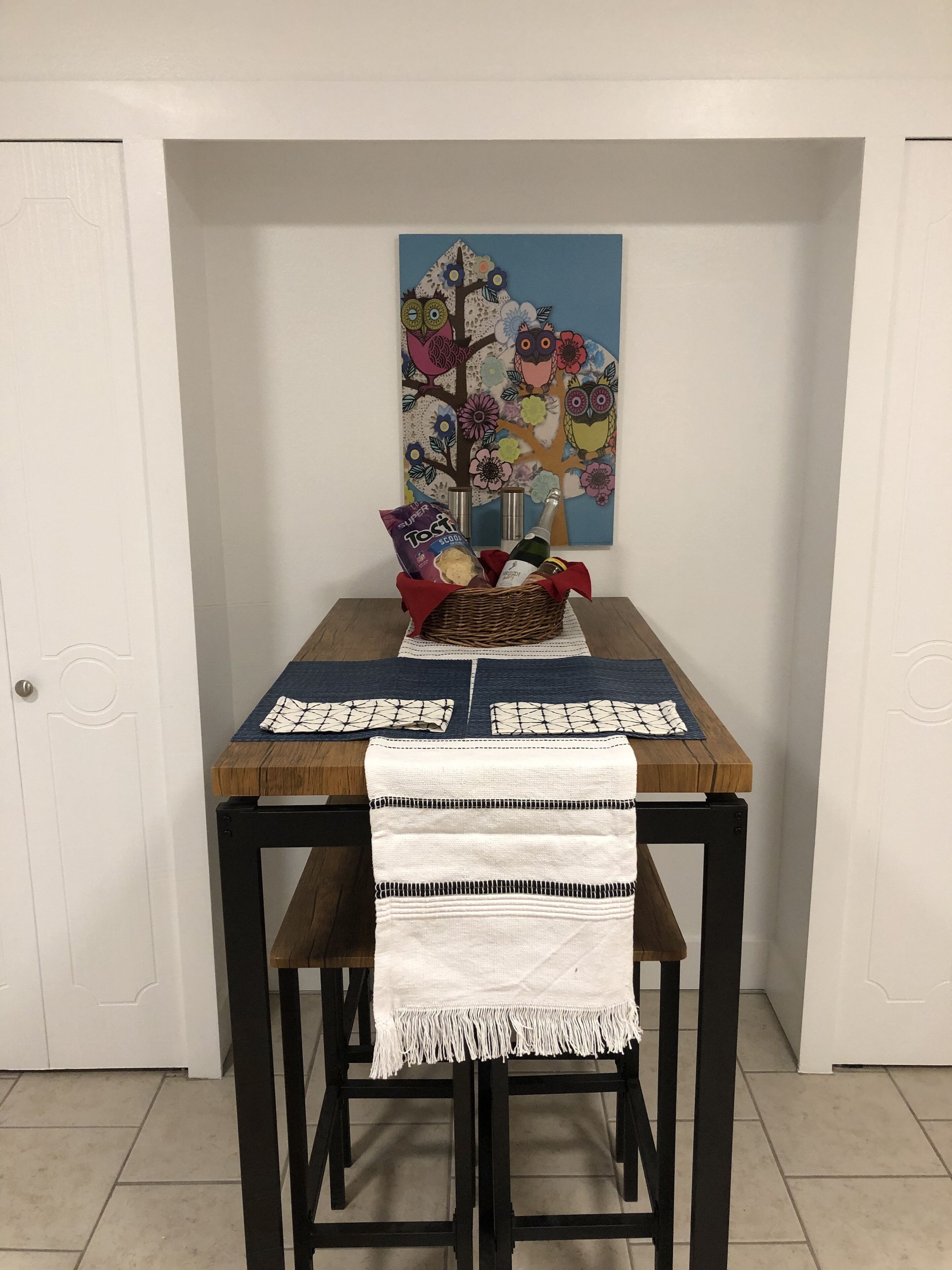 Une table à manger avec un set de table bleu, un chemin de table blanc et un panier en osier contenant des en-cas et une bouteille.