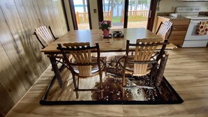 Une table de salle à manger en bois, quatre chaises, une horloge au mur et une vue sur l’extérieur par une porte.