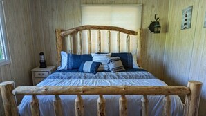 Un lit en bois, avec un couvre-lit blanc et des oreillers bleus et blancs, dans une pièce aux murs en bois et dotée d’une fenêtre.