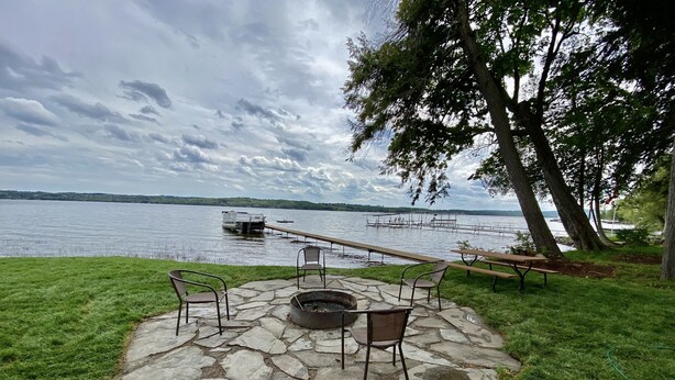 Un foyer en pierre, avec des chaises et une table de pique-nique, situé au bord d’un lac, entouré d’herbe et d’arbres.