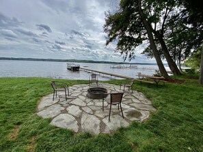 Un hogar de piedra con sillas y una mesa de picnic junto a un lago, rodeado de césped y árboles.