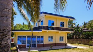 Uma casa amarela de dois andares com varanda, rodeada por um pequeno gramado e palmeiras.