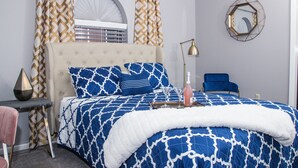 A bedroom with a tufted headboard, a bed with a blue and white patterned bedspread, a bedside table with a lamp, and a chair. - Modern Designer Condo Near I55 w/Balcony (Jackson)