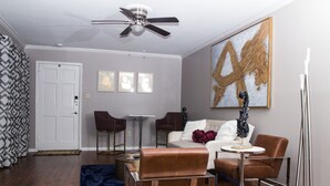 A modern living room with a sofa, two armchairs, a coffee table, and a floor lamp. There is a ceiling fan, a door, and a large abstract painting on the wall. - Modern Designer Condo Near I55 w/Balcony (Jackson)