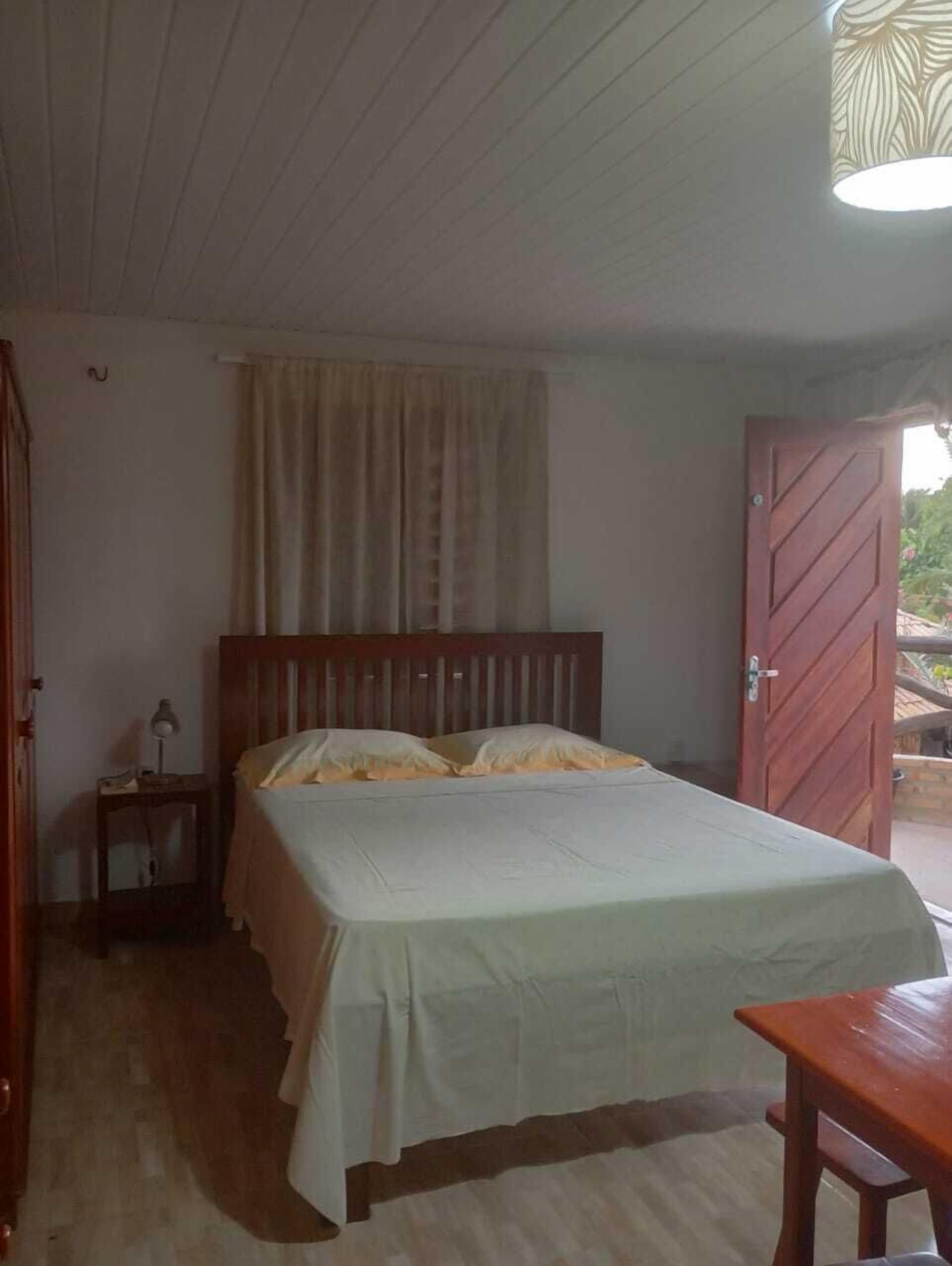 Un dormitorio con cama, mesita de noche, puerta de madera y ventana con cortinas.