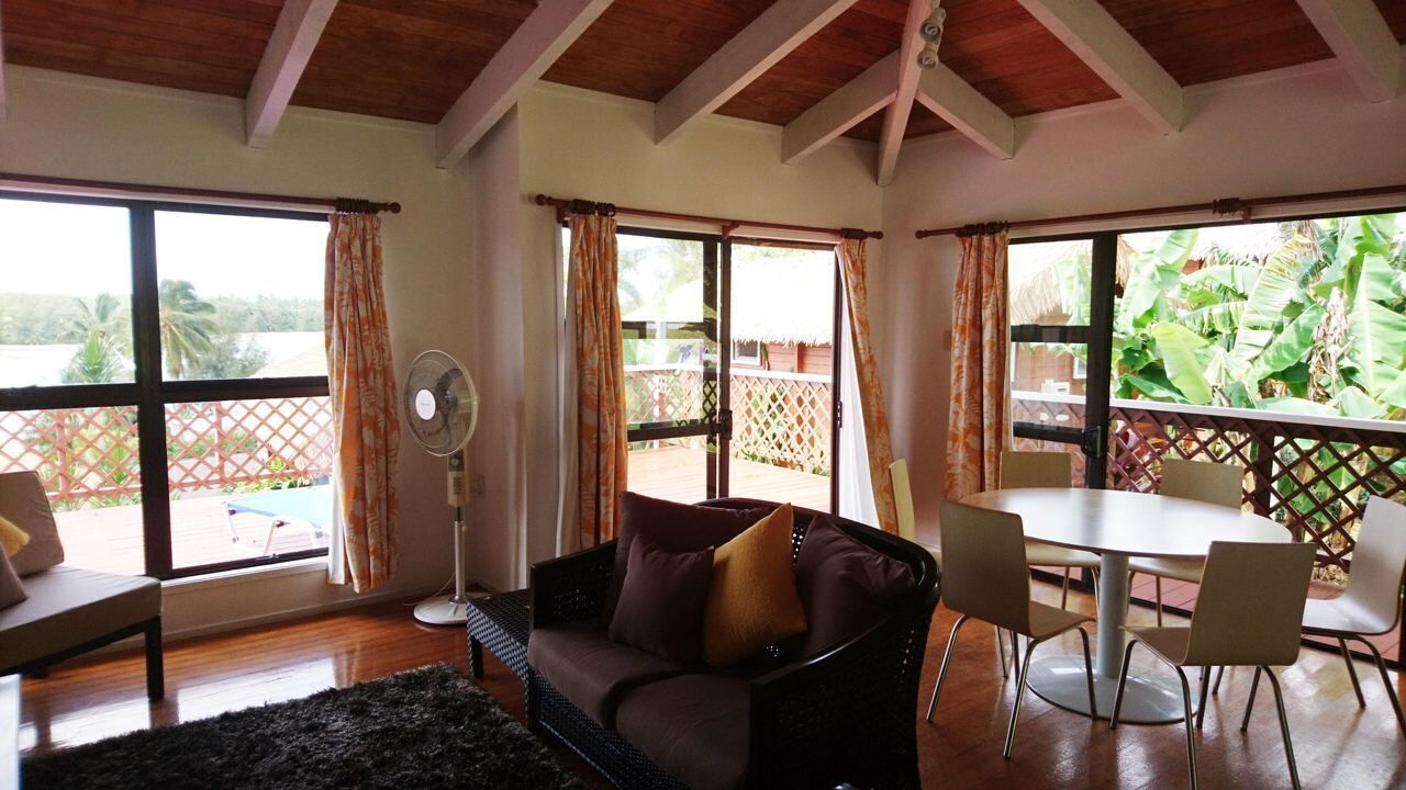 A room with a sofa, a dining table with chairs, a fan, and large windows with curtains.