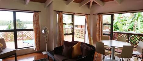 A room with a sofa, a dining table with chairs, a fan, and large windows with curtains.