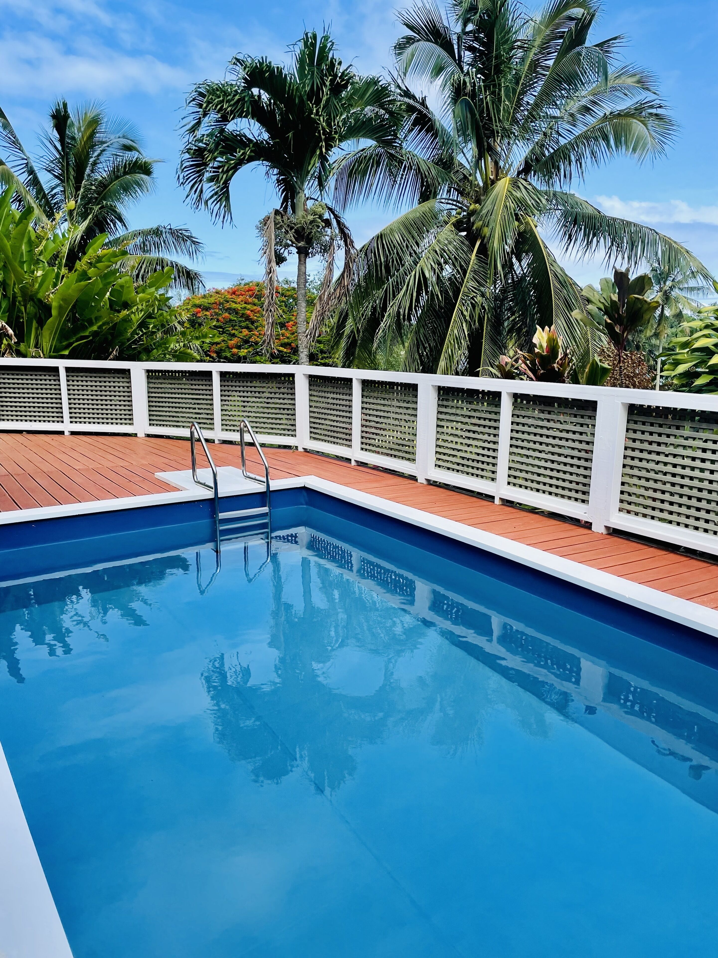 Piscina azul cristalina com deck branco e escada de metal, cercada por vegetação tropical exuberante e um cercado branco.