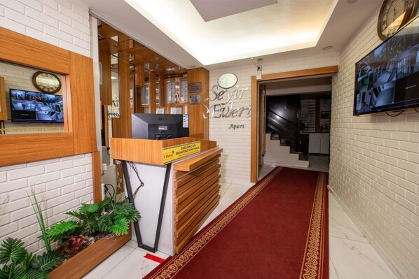 Zona de recepción con un mostrador de madera, un reloj, un televisor y una escalera que sube a otro piso.
