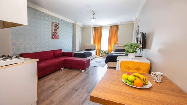 Un salon moderne avec un canapé rouge, une table à manger en bois sur laquelle on peut voir des fruits, et un coin cuisine avec une cuisinière.