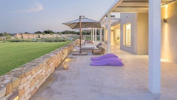 Een modern terras buiten, met een stenen muur, lounge stoelen en parasols.