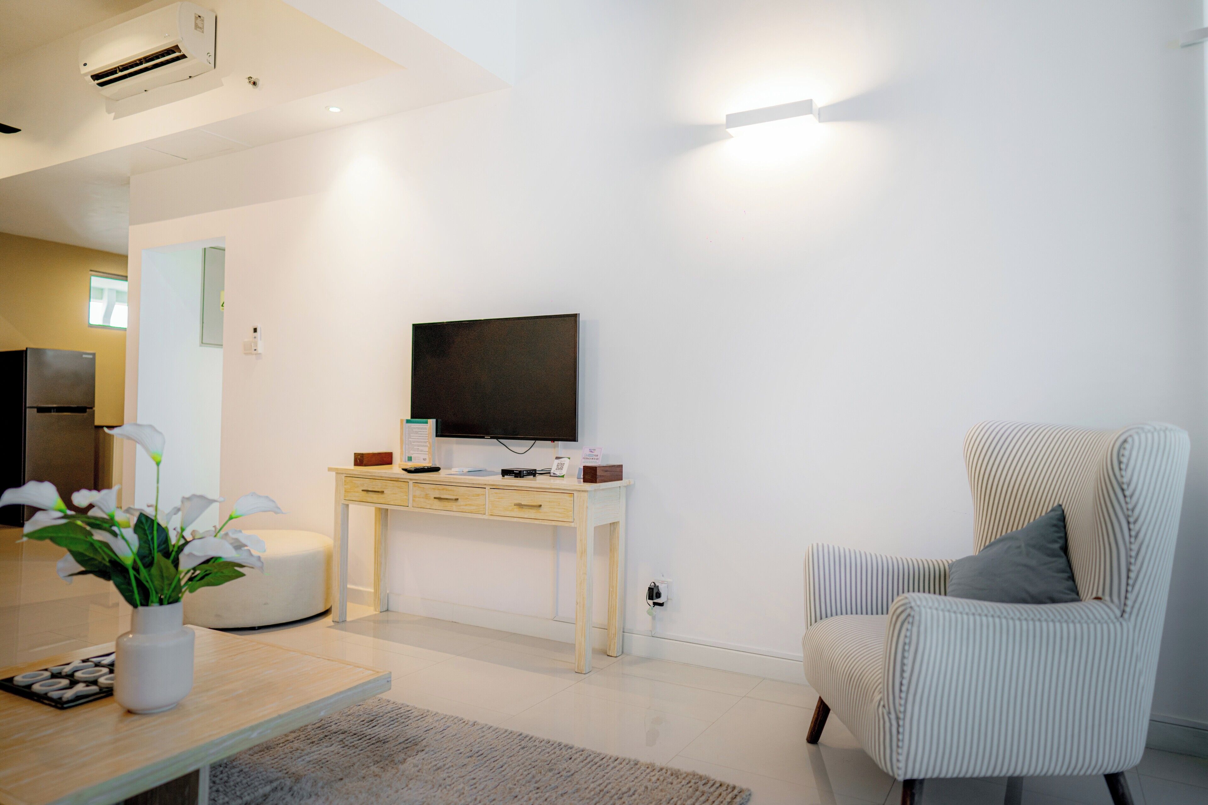 A modern living room with a flat-screen TV, a striped armchair, a small table with a vase of flowers, and a light-colored sofa.