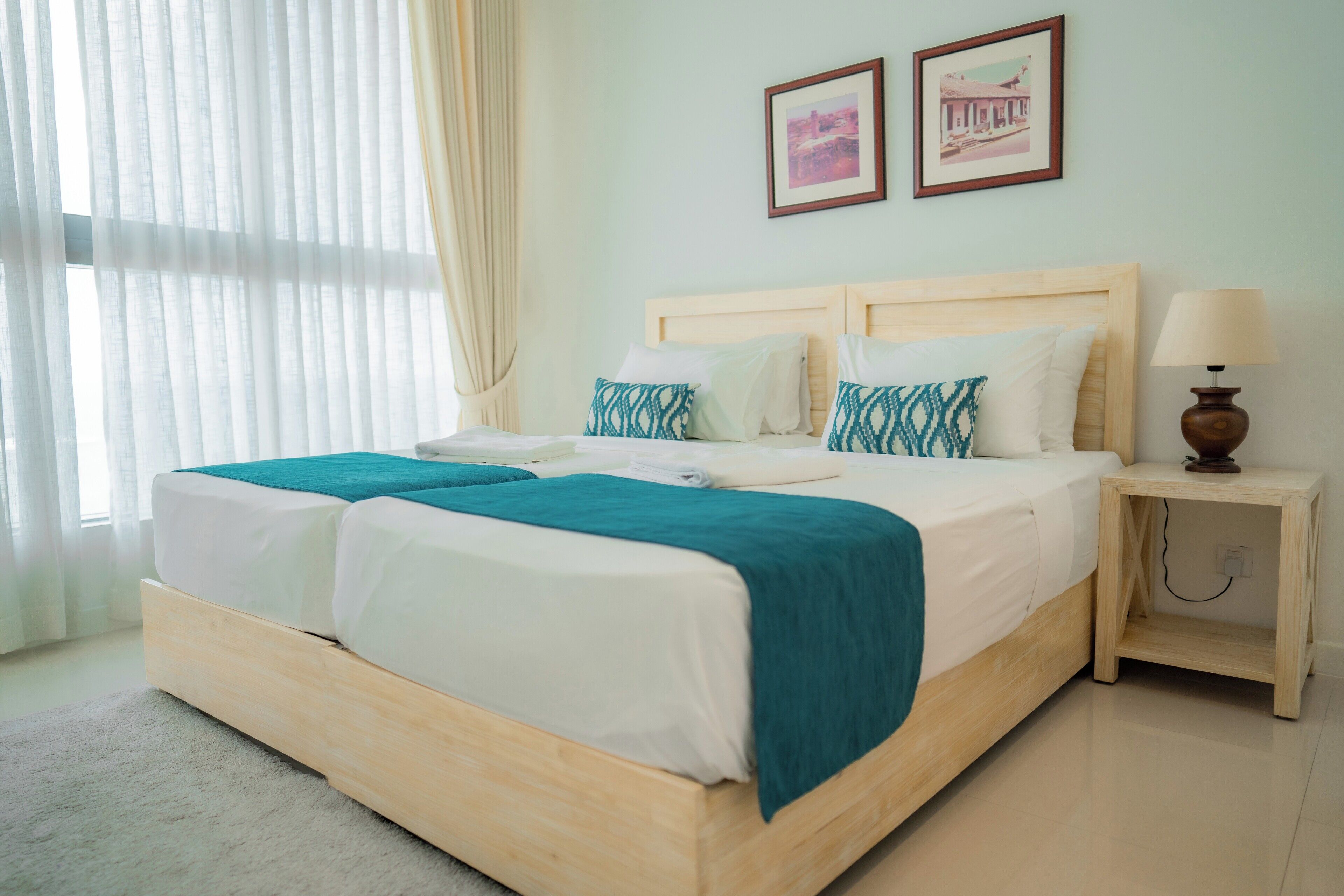 Een tweepersoonsbed met houten frame, wit beddengoed en turquoise sierkussens. Er staat een nachtkastje met een lamp en een stopcontact.