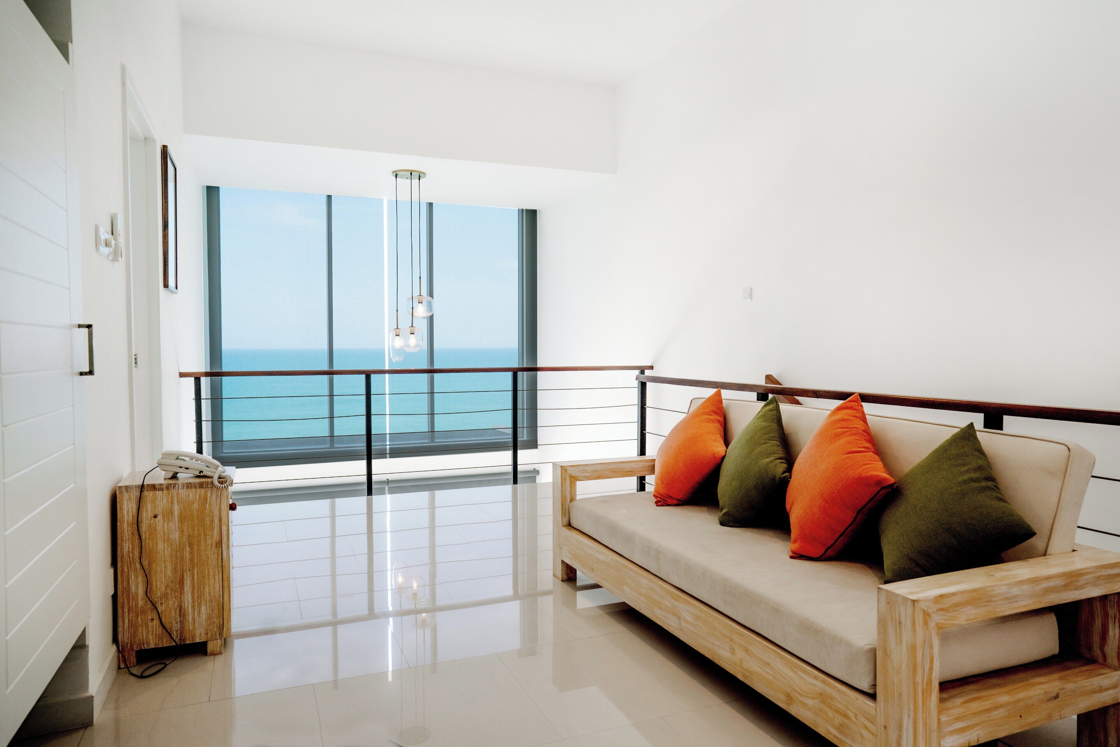 Una sala de estar moderna con un sofá, cojines de colores, una mesita de madera con un teléfono y un ventanal con vistas al mar.