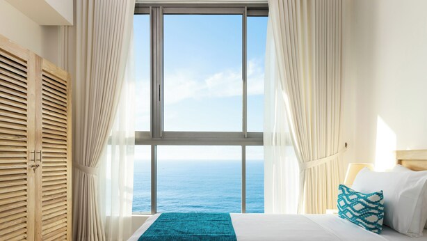 A bedroom with a large window, a bed with white and teal bedding, and a wooden wardrobe. - Oceanfront Galle by ICC (Galle)