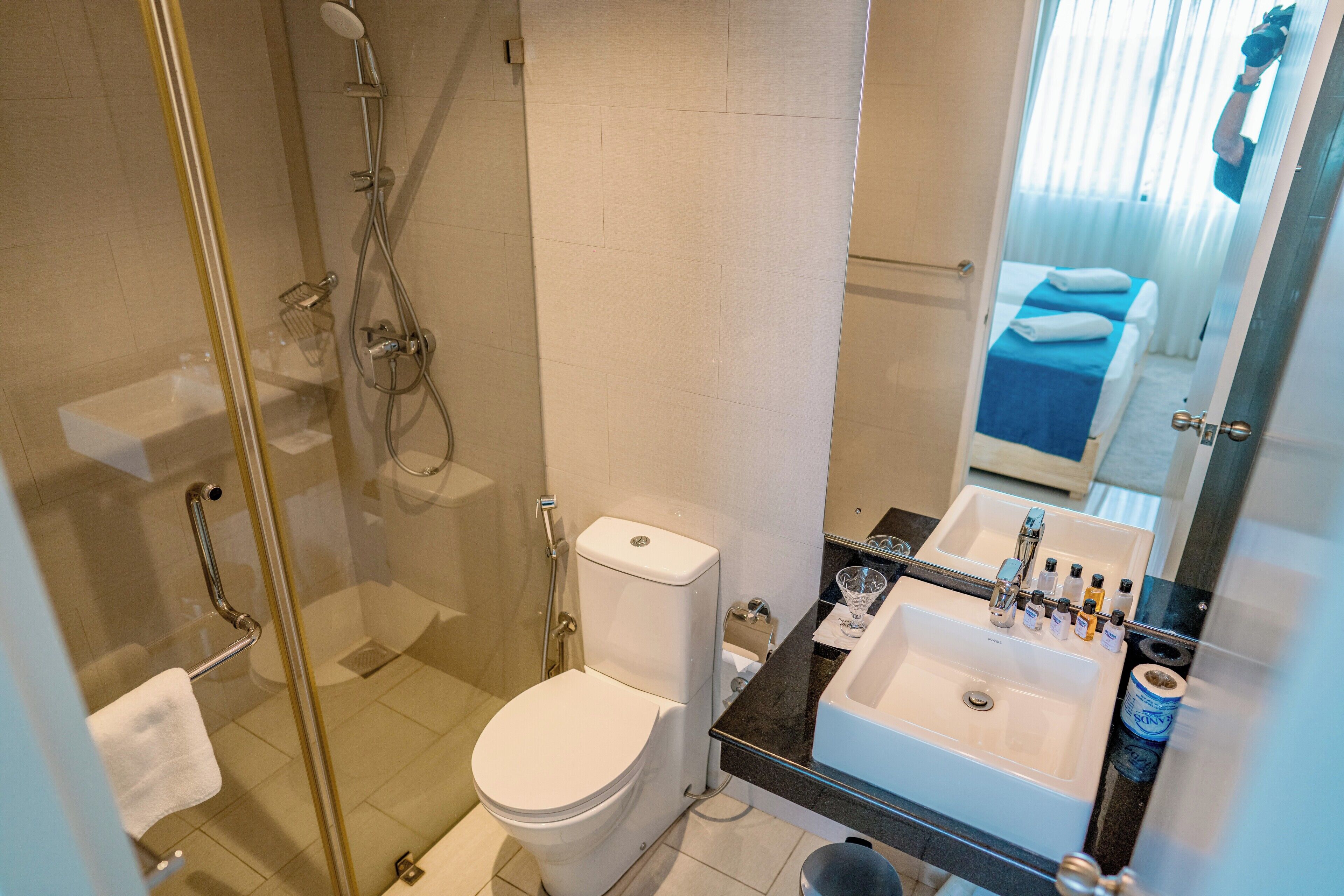 Un baño moderno con ducha de vidrio, inodoro blanco, lavamanos con artículos de aseo y un espejo que refleja una cama y una ventana.