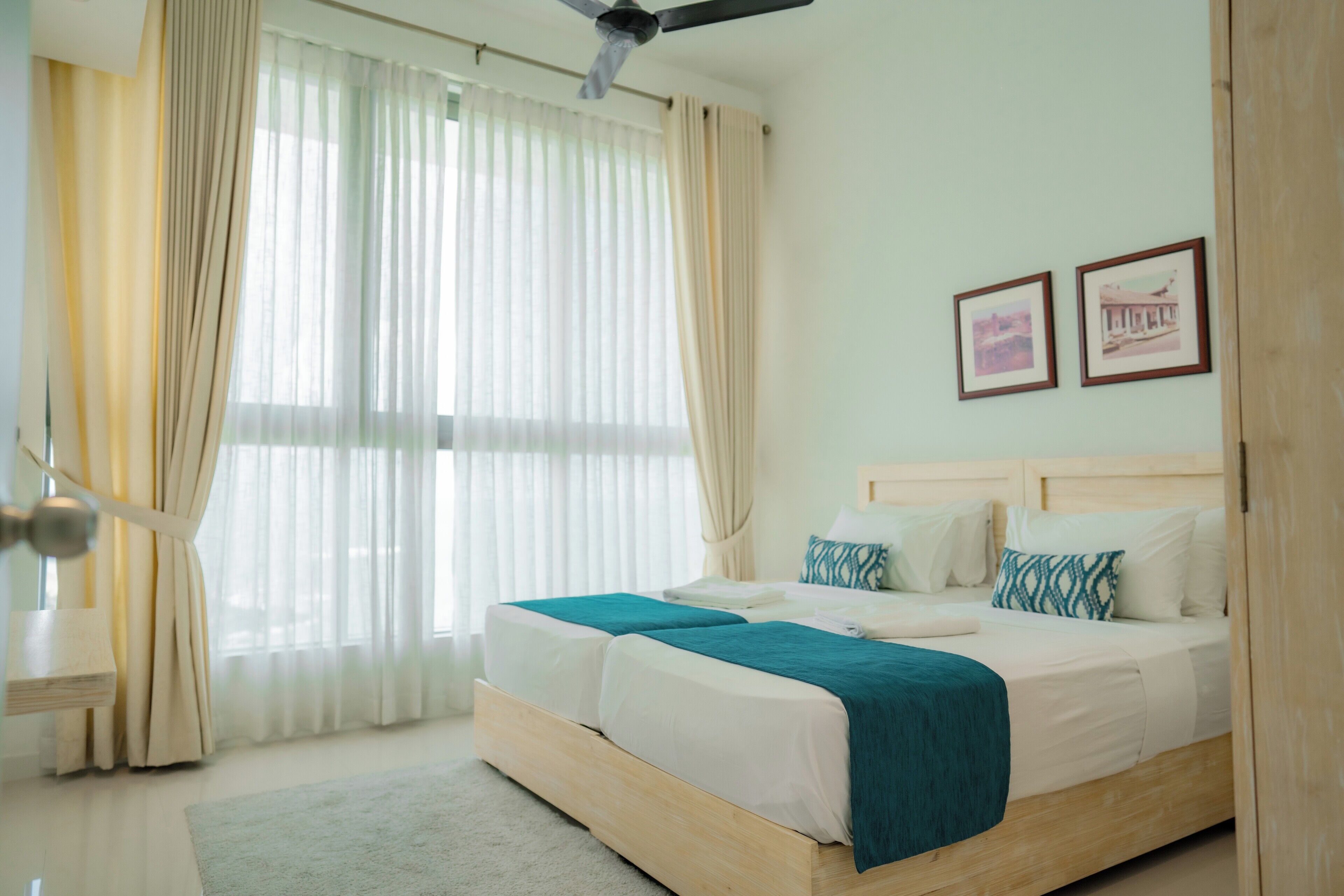A bedroom with a wooden bed, white bedding, and a teal throw blanket. There are two framed pictures on the wall and a ceiling fan.