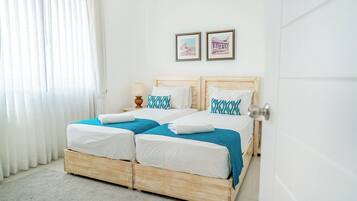 Una cama doble con ropa de cama blanca y azul, una mesita de noche con lámpara y dos cuadros en la pared.