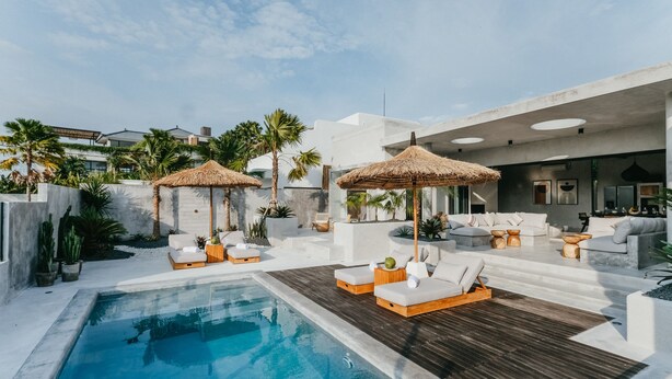 Un área de piscina moderna al aire libre con sillones de descanso y sombrillas de paja, rodeada de palmeras y un edificio blanco.