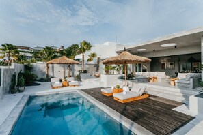 A modern outdoor pool area with lounge chairs and thatched umbrellas, surrounded by palm trees and a white building. - Concrete Retreat (Canggu)