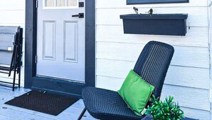 A front porch with a white door, a black chair with a green cushion, and a small potted plant.