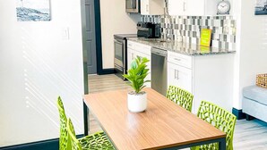 A modern kitchen with white cabinets, a microwave, and a sink. There is a dining area with a wooden table and green chairs.