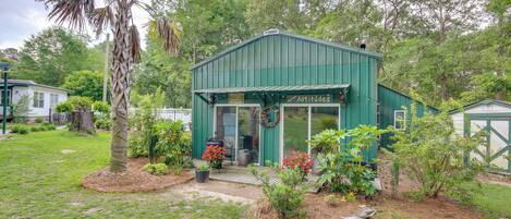 A green metal building with a covered porch, surrounded by a garden with various plants and a palm tree.
