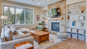 A modern living room with a stone fireplace, a large TV, and a bookshelf. - Winter Park Condo w/ Balcony ~ 4 Mi to Slopes (Winter Park)
