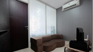 A modern living room with a sectional sofa, a flat-screen TV on a wooden stand, and a large window with vertical blinds. - Simply Clean And Cozy 1Br With Sofabed At Atlanta Residence Apartment (Depok)