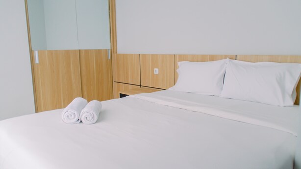 A neatly made bed with white linens and two rolled towels, a wooden headboard, and a small bedside table. - Elegant And Comfy 2Br Transpark Bintaro Apartment (South Tangerang)