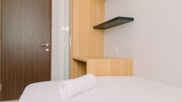 A hotel room with a wooden door, a white bed with a rolled-up white towel, and a wooden shelf unit against the wall.