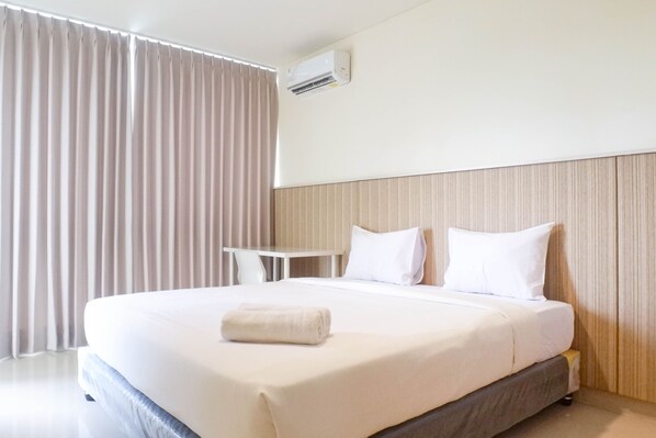 Quarto de hotel com uma cama grande, roupa de cama branca, uma mesinha e uma janela com cortinas.