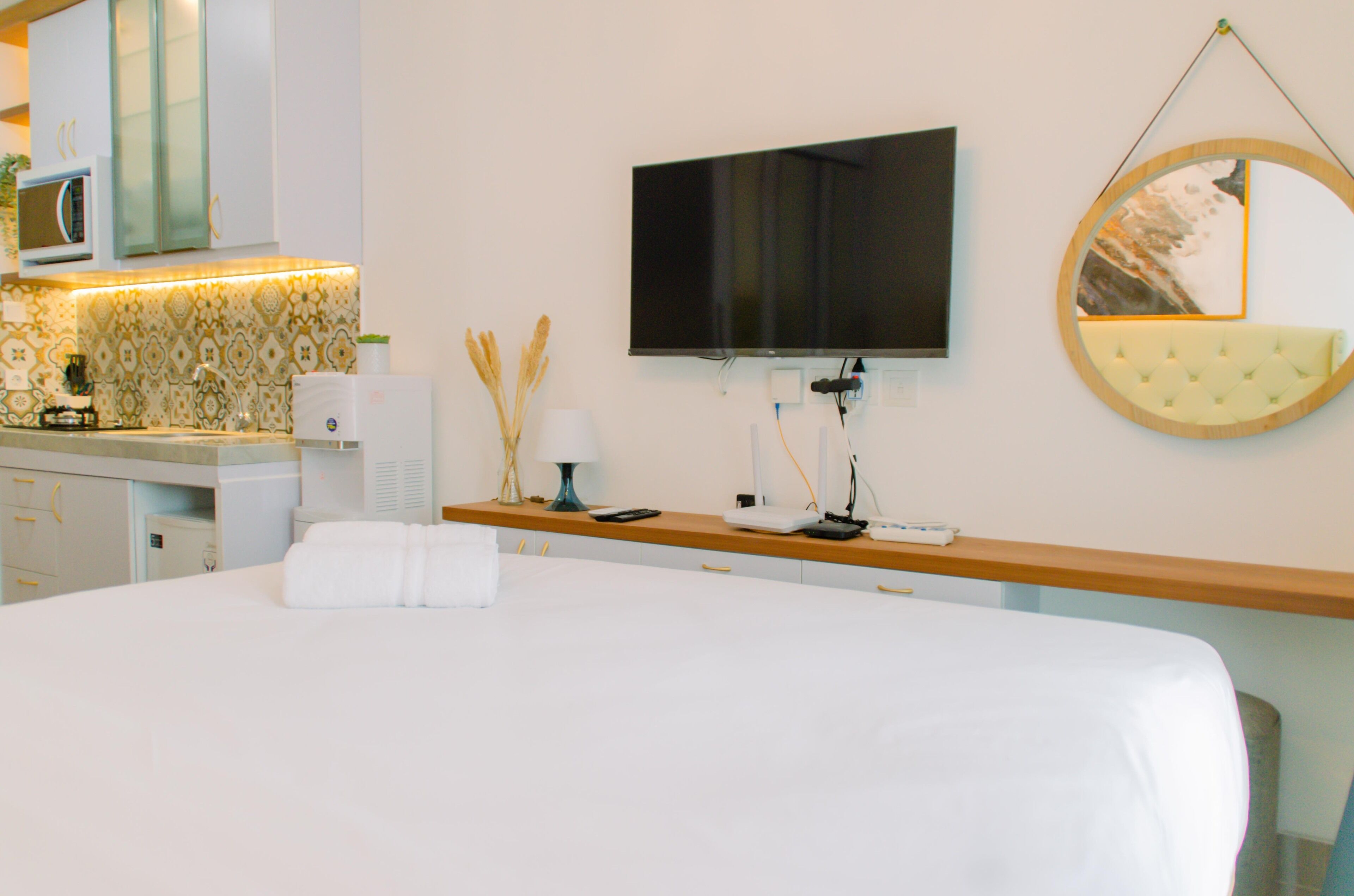 Habitación de hotel con cama, televisor montado en la pared, espejo redondo y mesita de noche de madera con lámpara y control remoto.