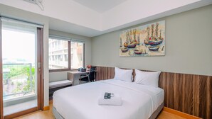 A hotel room with a large bed, a desk with a chair, a window with a view, and a painting of sailboats on the wall. - Comfortable Pool View Studio Room at LRT City Jatibening Apartment (Bekasi)