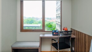 A small room with a desk, a chair, a bench, and a window with a view of buildings and greenery. - Comfortable Pool View Studio Room at LRT City Jatibening Apartment (Bekasi)