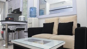 Un soggiorno moderno con un tavolino in vetro, un divano beige con cuscini neri, angolo cottura con mobili bianchi, un dispenser d'acqua e uno sgabello da bar.