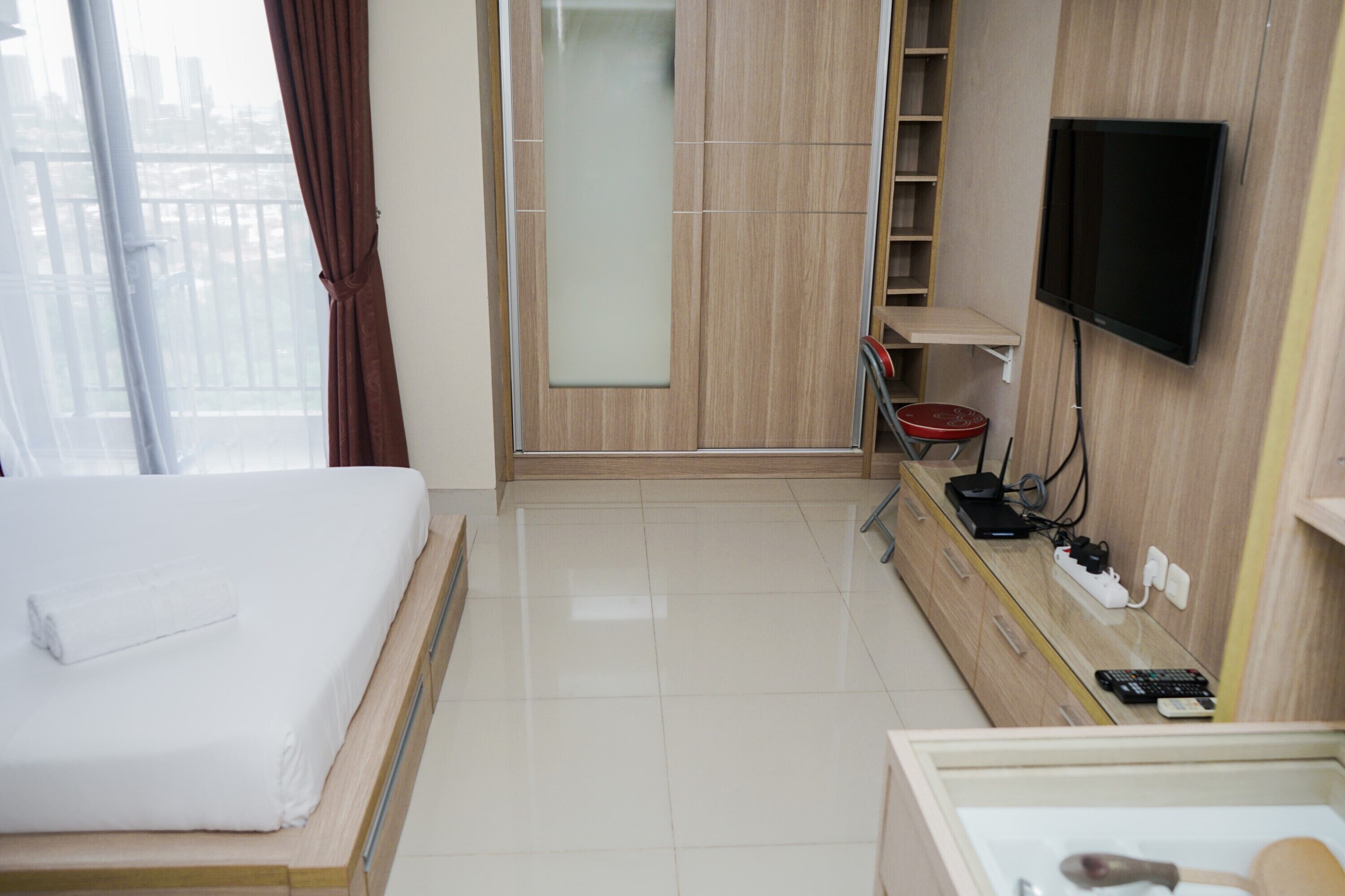 A hotel room with a bed, a TV mounted on the wall, a desk with a computer and phone, and a window with curtains.