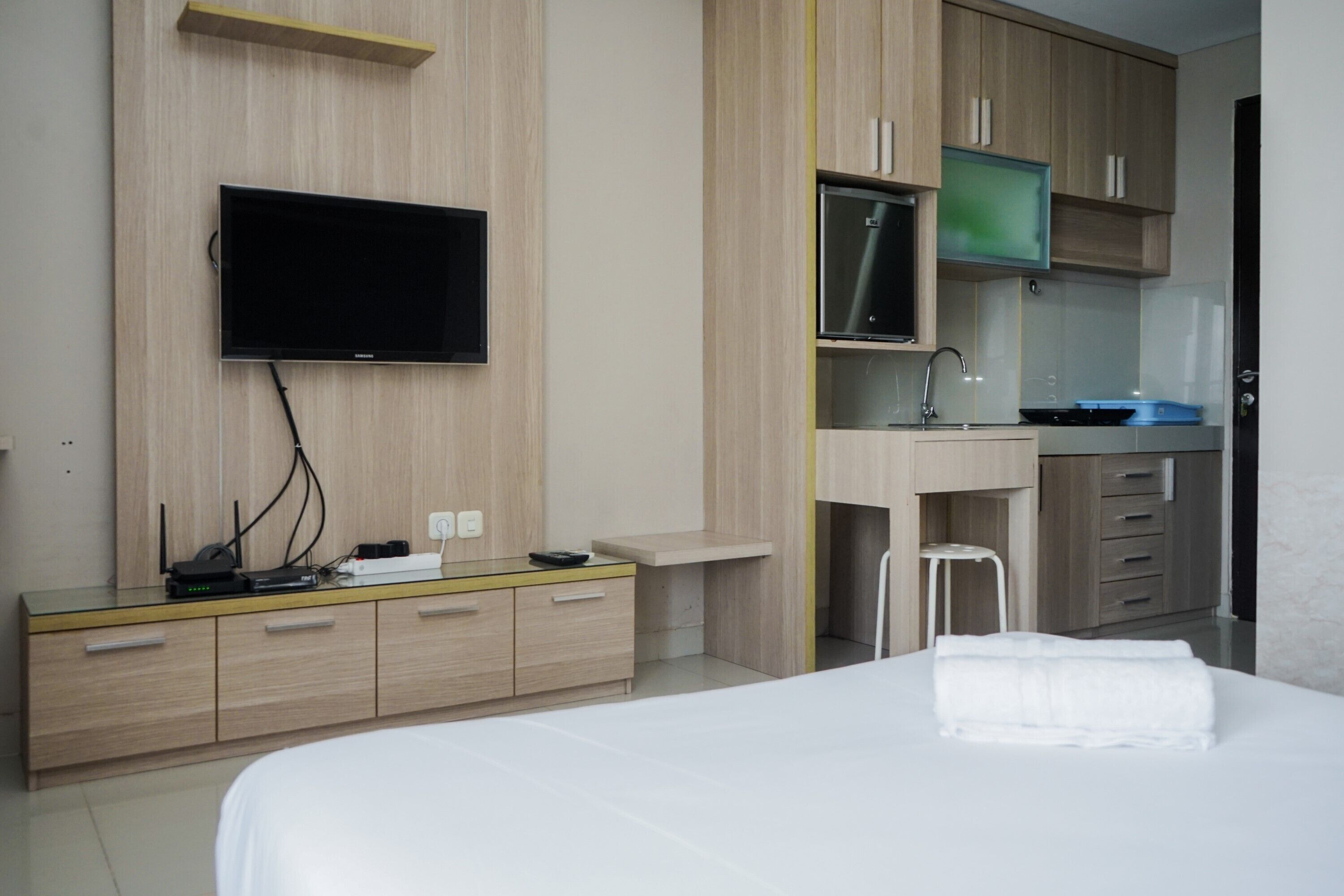 A hotel room with a bed, a TV mounted on the wall, a desk with a sink, and a small stool.