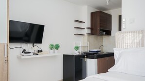 A compact hotel room with a bed, a TV mounted on the wall, a small kitchenette, and a shelf with potted plants. - Comfy And Minimalist Studio Room At Serpong Garden Apartment (Cisauk)