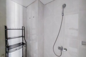 A bathroom with white tiled walls, a shower with a handheld hose, and a metal shelf unit with a white basin. - Comfy And Minimalist Studio Room At Serpong Garden Apartment (Cisauk)