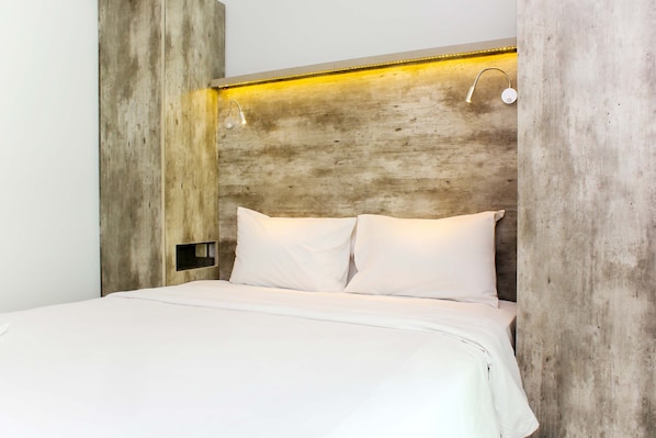 Una cama con ropa blanca, cabecera de madera y dos lámparas fijadas a la pared.