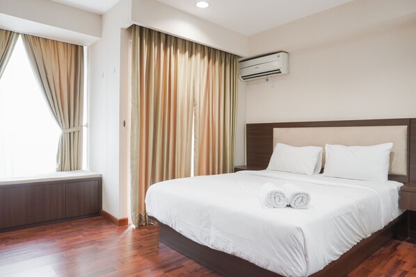 A hotel room with a large bed, a window with curtains, an air conditioning unit, and a wooden floor.